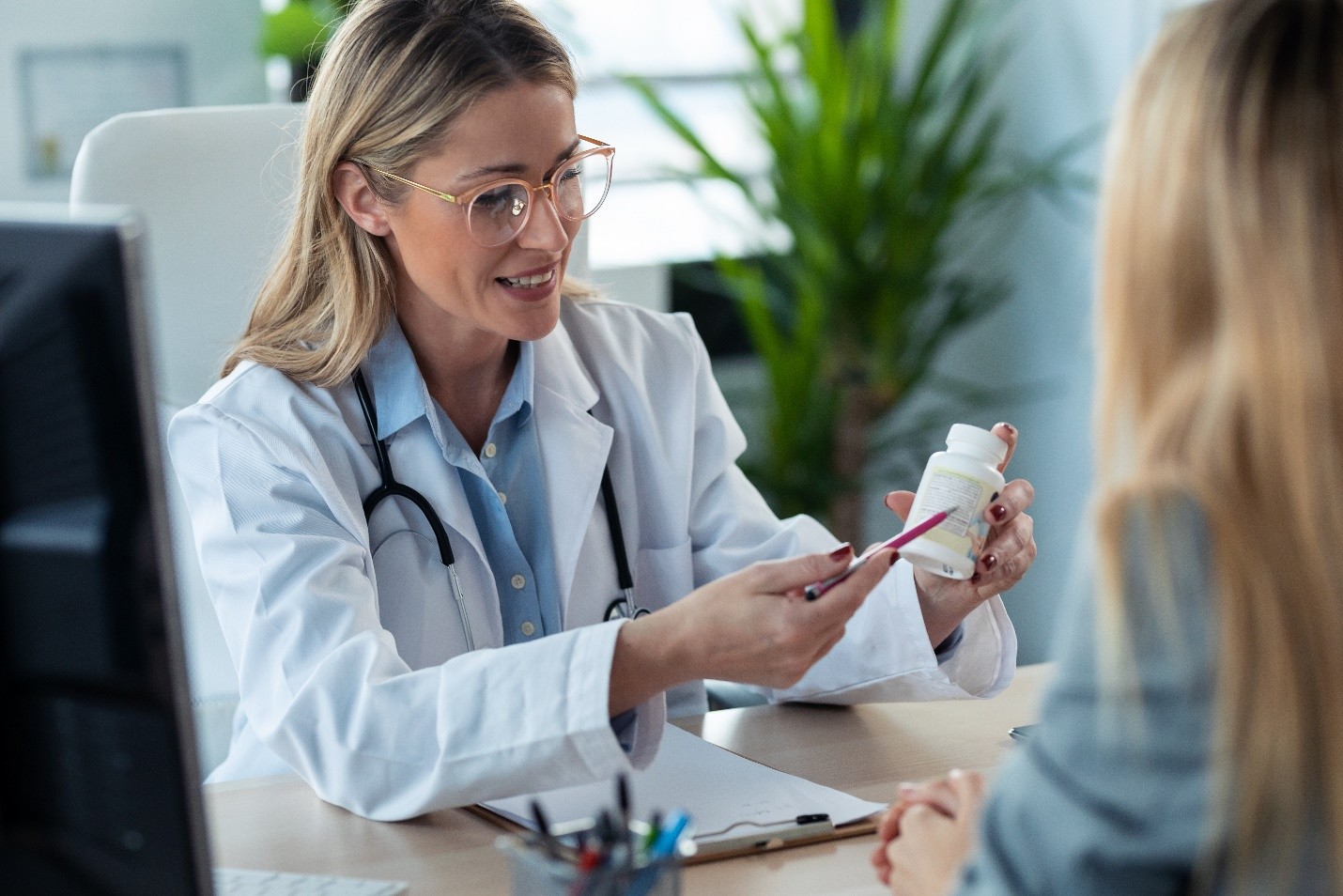 A female doctor holding a white vitamin bottle, explaining the details of its nutritional panel to patient in a consultation.